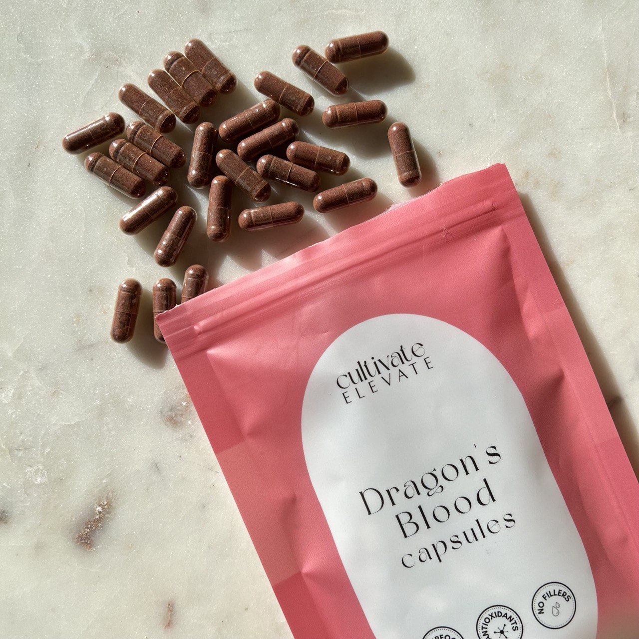 Pink package of 'Dragon's Blood capsules' by Cultivate Elevate on a marble surface with scattered capsules.