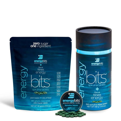 A small canister of ENERGYbits Spirulina next to its packaging, with several tablets visible in front. The canister and packaging are blue with product information and branding.